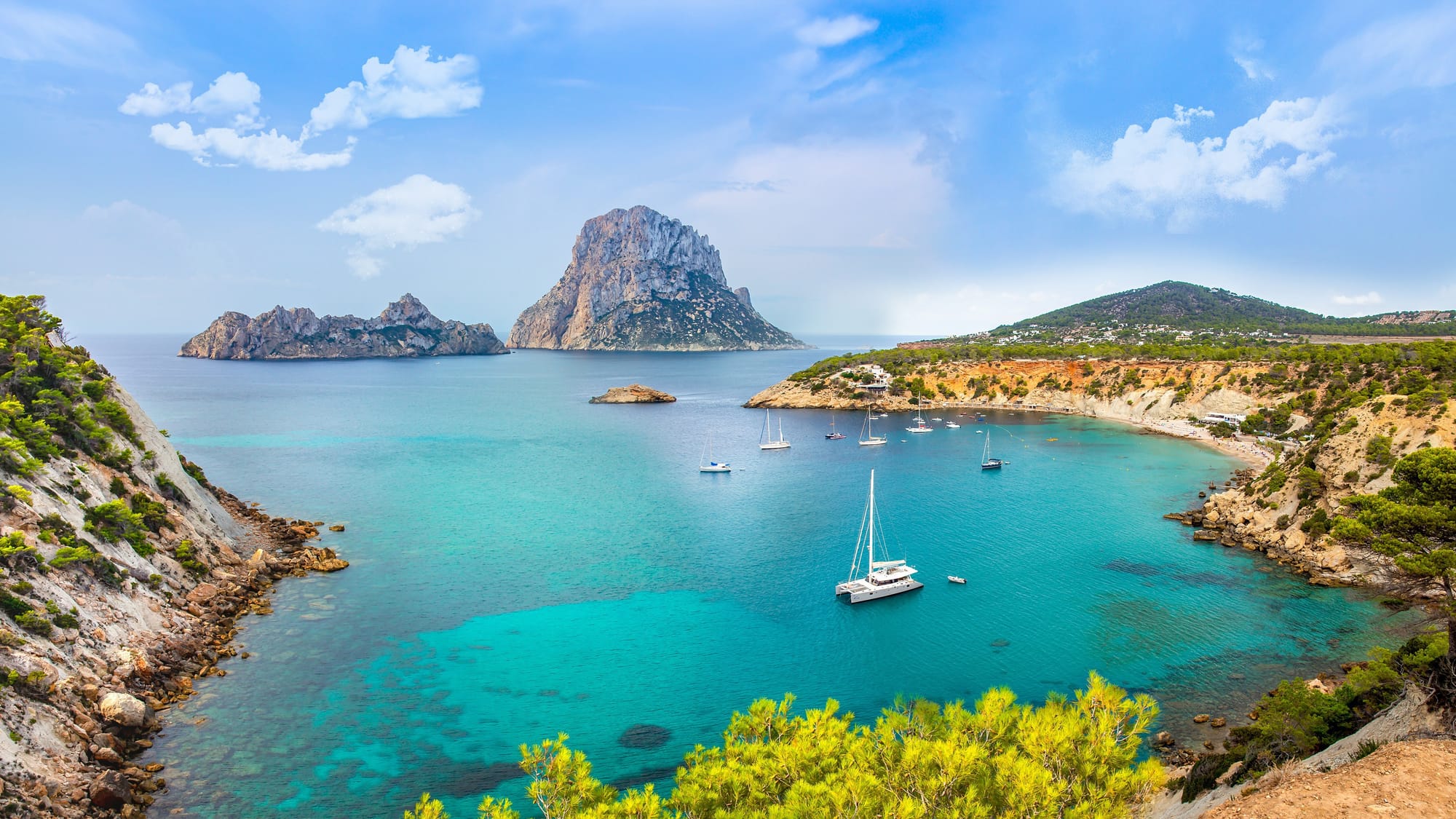 Scenic coastal view of Ibiza with turquoise waters, rocky cliffs, and sailboats anchored in a calm bay, with the towering islet of Es Vedrà in the background under a bright blue sky.