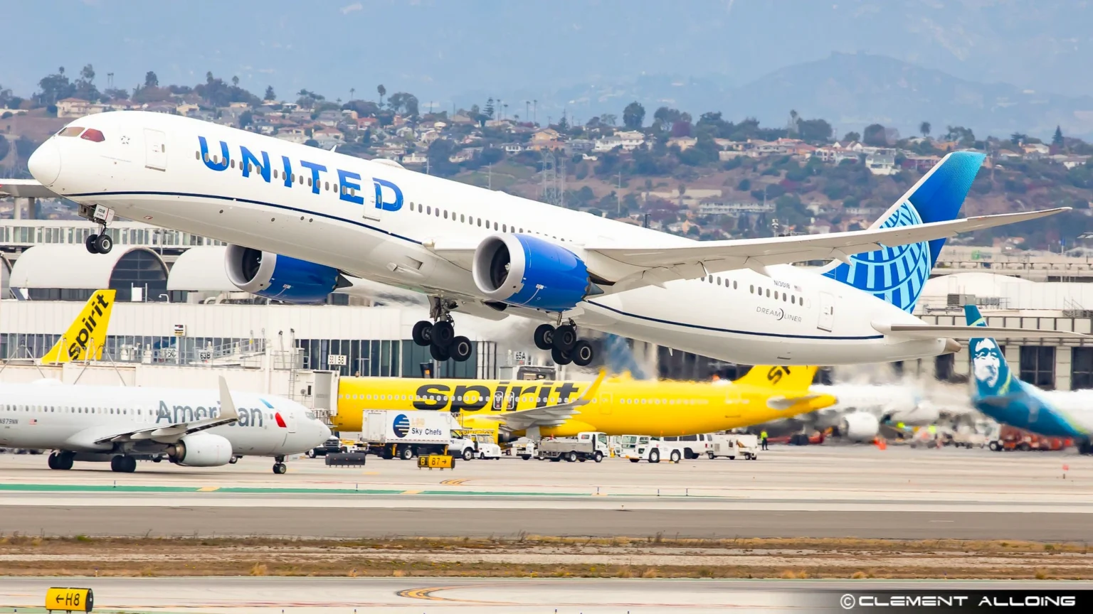 A United Airlines aircraft taking off from a busy airport runway, with planes from Spirit Airlines, American Airlines, and Alaska Airlines parked in the background.