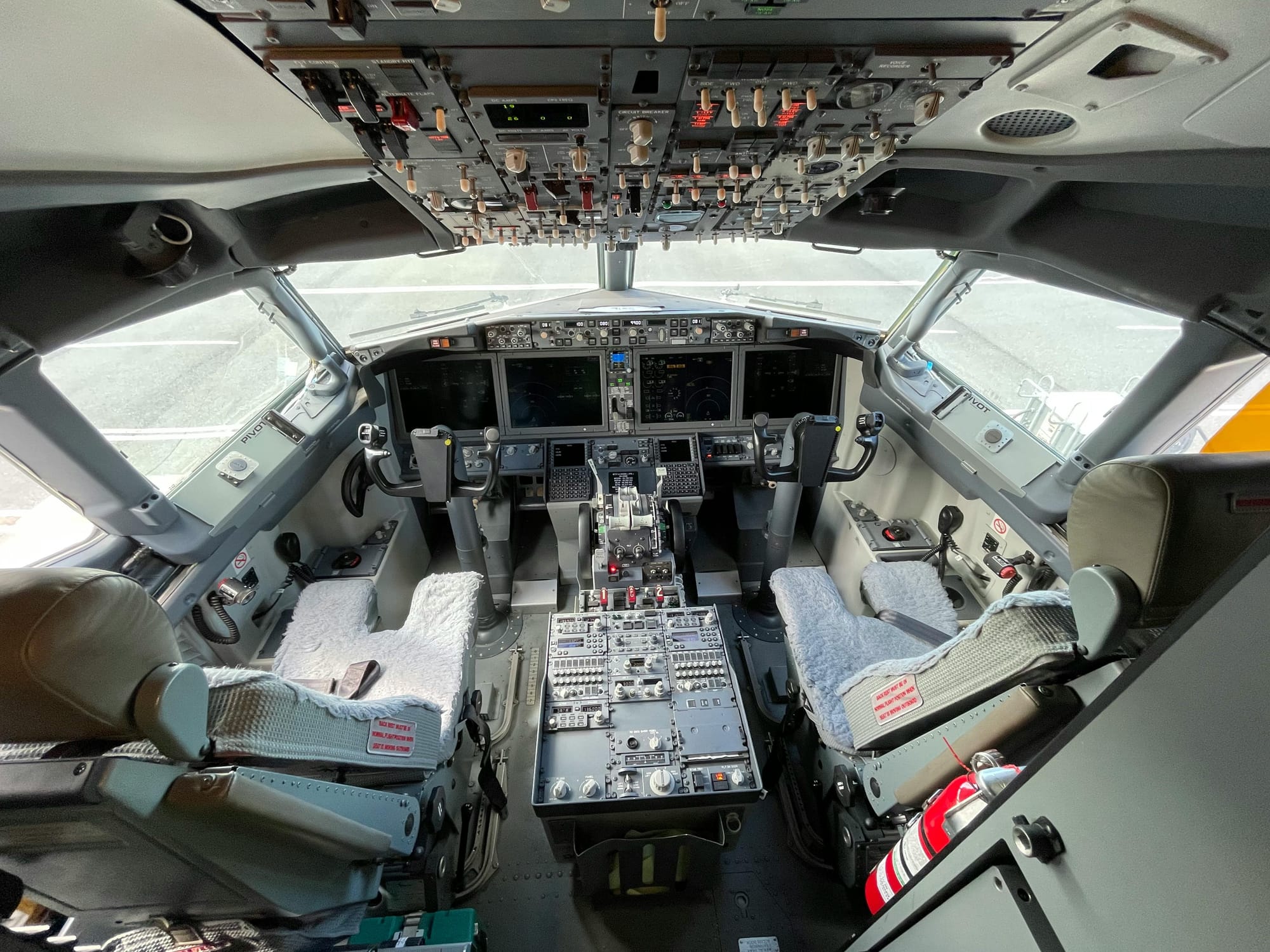  Cockpit view of a modern aircraft featuring two control yokes, multiple digital instrument displays, and various switches and levers, with textured seat coverings visible in the foreground.