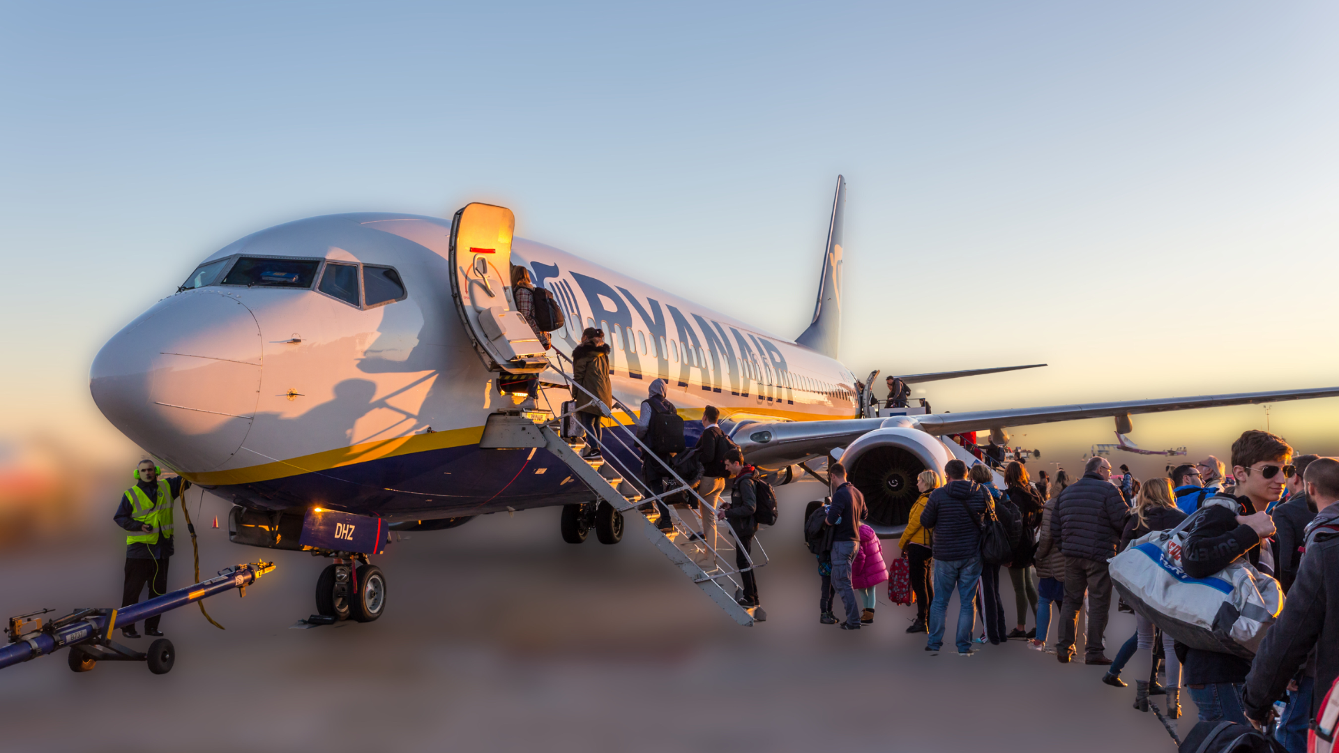 Passengers boarding a Ryanair aircraft from the tarmac via stairs, with a long line of travelers carrying bags. The scene is during sunset, casting a warm glow on the plane and people.