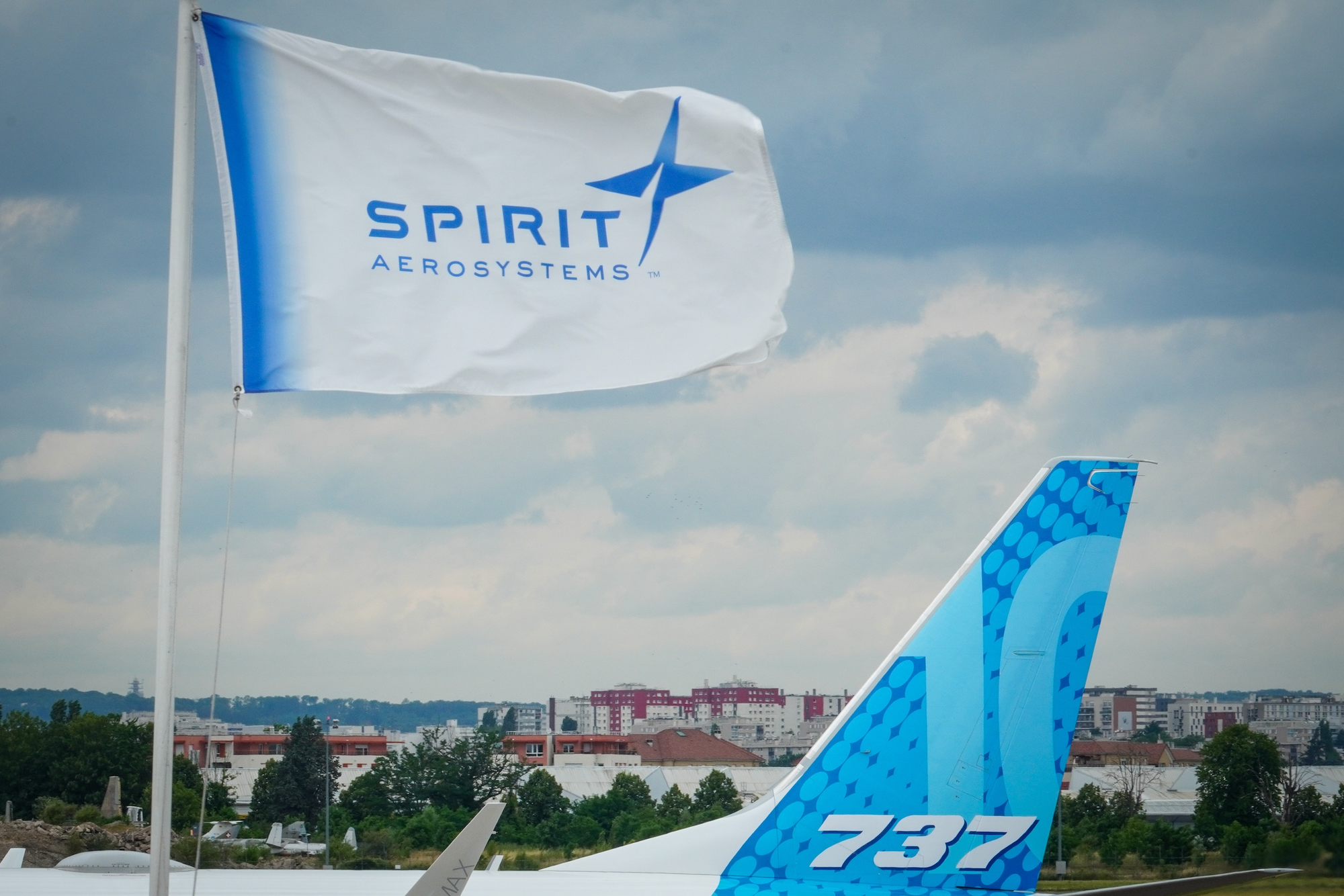 A Spirit AeroSystems flag waves in the foreground, while the tail of a Boeing 737 MAX aircraft is visible in the background. The image highlights the connection between Spirit AeroSystems and Boeing’s aircraft production.