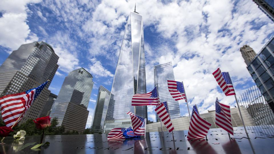 Photo of 9/11 Memorial with several American flags in the foreground and Skyscrapers in the background.