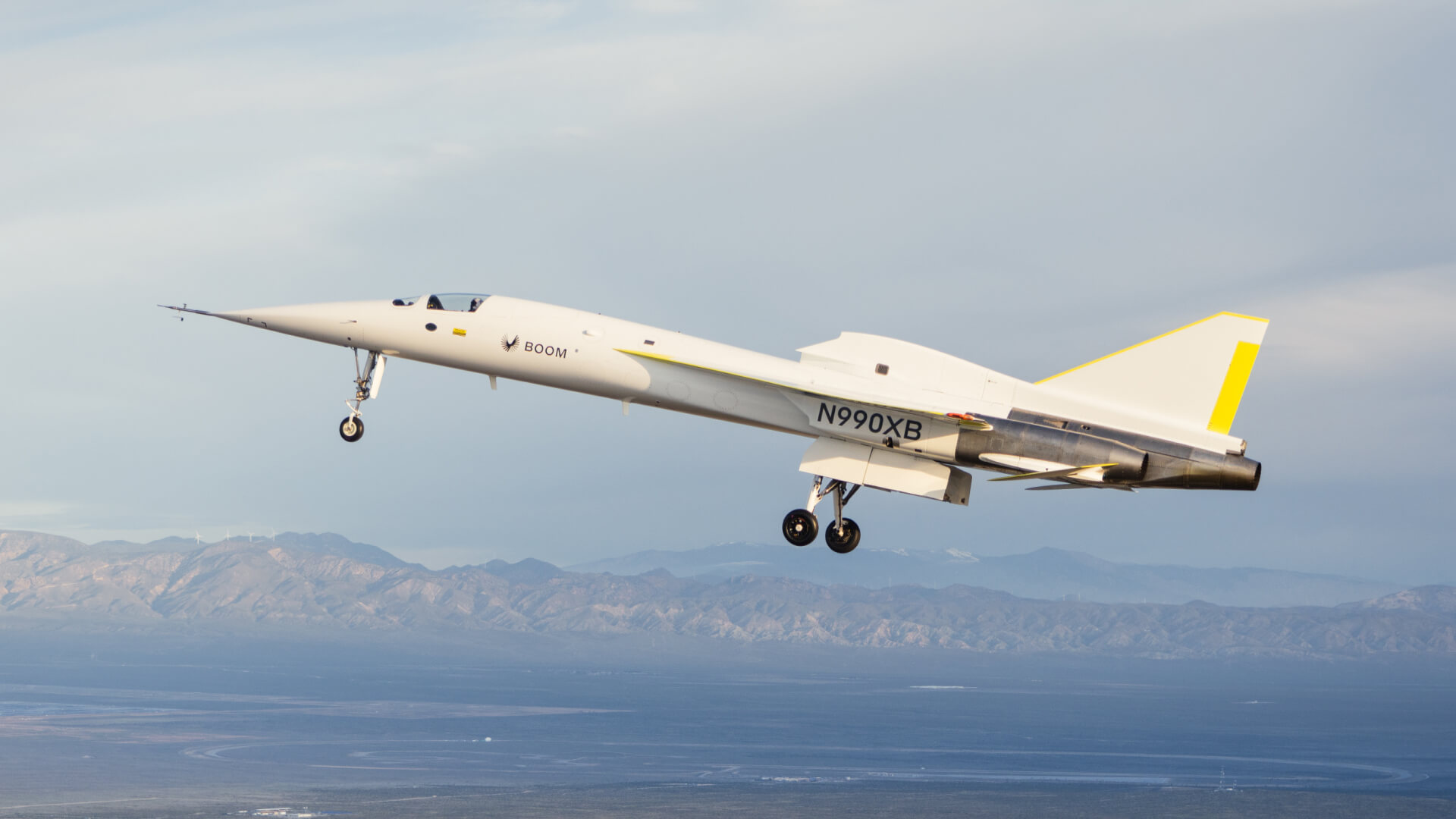 A sleek, white Boom Supersonic XB-1 demonstrator aircraft with yellow accents and landing gear extended.