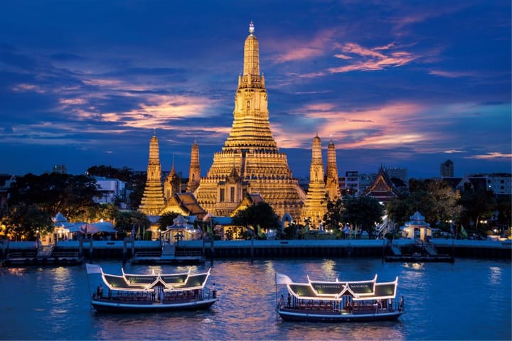 Wat Arun (Temple of Dawn) illuminated at sunset with boats on the Chao Phraya River in Bangkok, Thailand.
