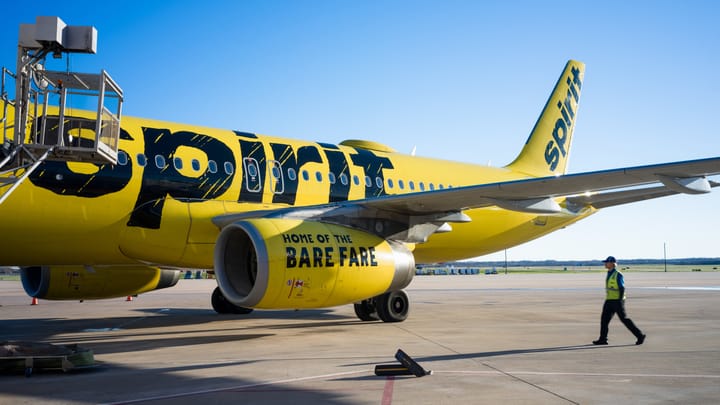 Spirit Airlines bright-yellow airplane on the tarmac, showing the 'Home of the Bare Fare' slogan, with ground crew walking nearby.