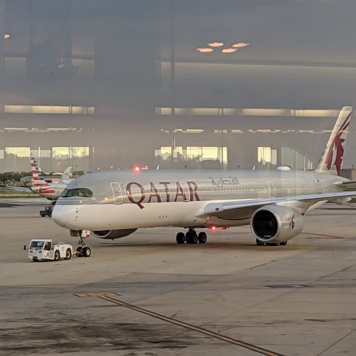 Qatar Airways aircraft on the tarmac at an airport, with an American Airlines plane visible in the background.